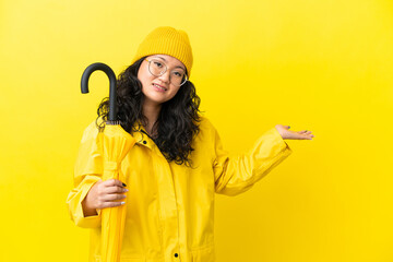 Asian woman with rainproof coat and umbrella isolated on yellow background extending hands to the...