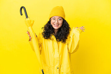 Asian woman with rainproof coat and umbrella isolated on yellow background with shocked facial...