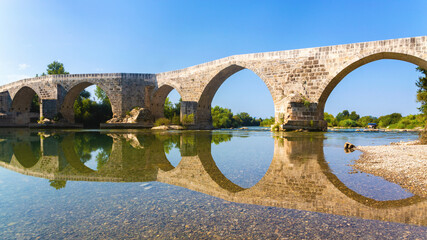 Fototapeta premium Historic Selcuk bridge in Aspendos reflects on the tranquil river, a picturesque scene for cultural tourism. Manavgat, Antalya, Turkiye (Turkey)