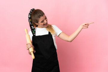 Young blonde woman holding a rolling pin isolated on pink background pointing finger to the side and presenting a product