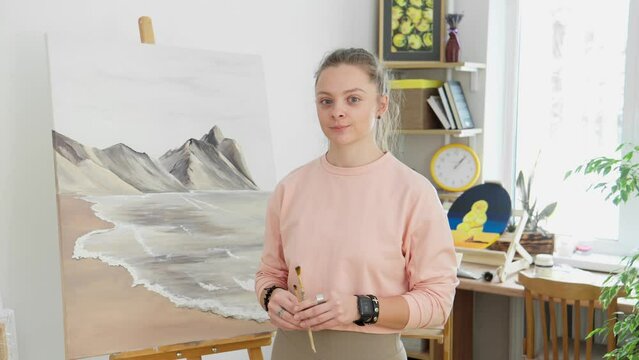 Portrait of talented young female artist looking at camera with artwork at background. Woman crosses arms while holding brushes. Talented woman artist indoors. Drawing concept. 4K, UHD