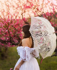 girl bride in vintage dress with white lace umbrella in park near blossoming tree with pink flowers