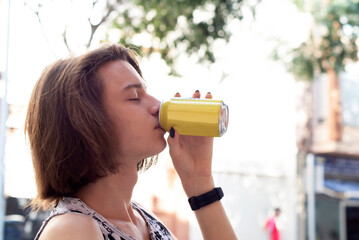 A teenage boy with long hair drinks gas water from a can, an energy drink on a hot day on the street