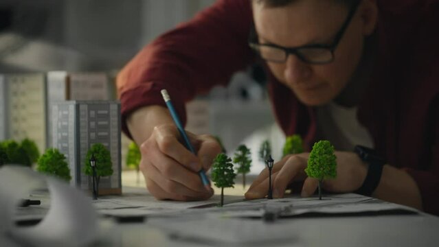 While working in the design studio of an industrial enterprise, design engineer, in his modern shirt, carefully applies a pencil to develop blueprints of green residential area, showing his creativity