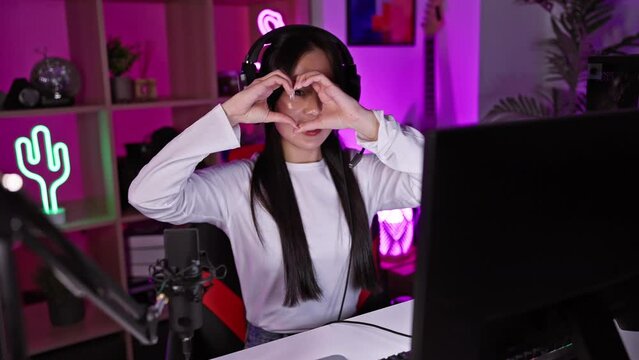 A cheerful young asian woman making a heart shape with her hands in a vibrant gaming room with neon lights at night.