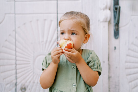 Little girl gnaws a red apple while standing near a white carved wooden door and looks away