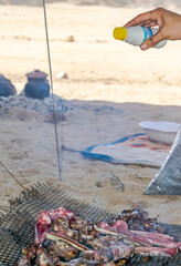 A makeshift barbecue outside in the Sahara desert, a hand holds a salt shaker and seasoning meat chops on a grill over a wood and coal fire. A clay pot casserole dish in blurred background on sunlight