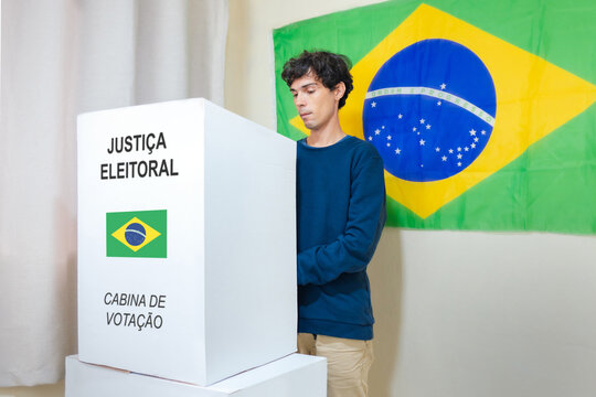 Jovem homem brasileiro escolhendo o seu candidato na urna eletrônica. Na parede, a bandeira do Brasil decora o local de votação. A imagem retrata as eleições brasileiras