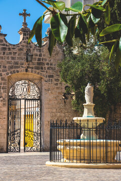 A quaint and peaceful courtyard, with a statue of the Virgin Mary and a little fountain as well as an ornate iron gate in the background. Location, Mellieha - Malta.