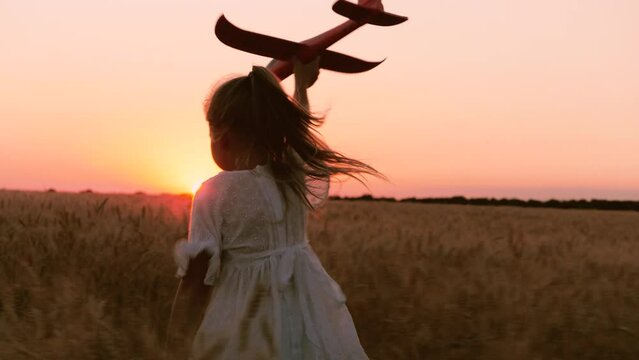 Happy little girl kid in white dress running with plane toy on wheat field at sunset sunrise horizon back view closeup. Adorable female child playing aircraft imagine pilot flying at dusk dawn meadow