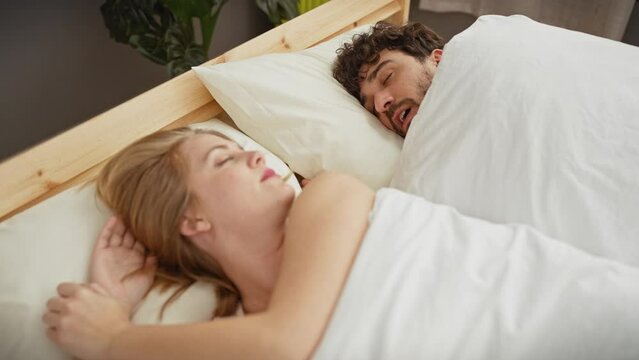 Woman frustrated in bed next to snoring man in a home bedroom setting, depicting couple, sleep, and relationship issues.