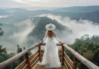 A woman in a white dress and hat standing on a wooden bridge, overlooking the misty forest landscape from a high altitude, with a panoramic view of mountains.