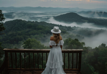 A woman in a white dress and hat standing on a wooden bridge, overlooking the misty forest landscape from a high altitude, with a panoramic view of mountains.