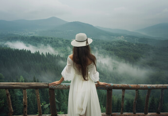 A woman in a white dress and hat standing on a wooden bridge, overlooking the misty forest...