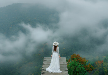 A woman in a white dress and hat standing on a wooden bridge, overlooking the misty forest landscape from a high altitude, with a panoramic view of mountains.