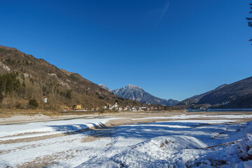 Empty frozen reservoir lake during winter time with village in the mountain landscape of the Alps,...