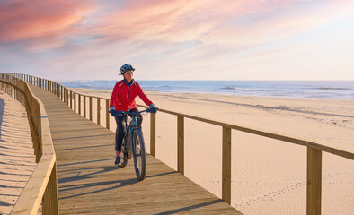happy active senior woman cycling during moody golden hour at the beach of the atlantic coast of...