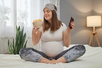 Poster Funny smiling pregnant woman sitting in lotus pose holding bowl with potato chips and bar of chocolate smelling harmful fast food braking pregnant diet © sementsova321