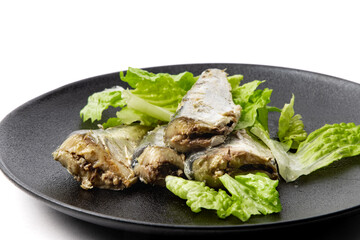 A black plate of tinned sardines garnished with pieces of lettuce isolated on white