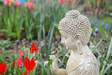 Portrait of a white garden buddha with his hand pointed up combined with red tulips. The hand gesture of this Buddha is called Abhaya Mudra