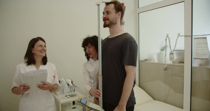 A doctor and a nurse measuring patient's height using stadiometer and modern, sunlit office in the hospital. They are working in positive atmosphere, having fun conversation and smiling.