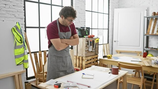 A young man with glasses and a beard, wearing a grey apron, appears thoughtful in a bright, windowed carpentry workshop.