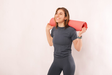 Young smiling woman in sportswear is holding a pink fitness mat over her shoulders.