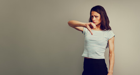 Beautiful fun casual grimacing woman in strip t-shirt showing deslike sign by hand down demonstrated negative signal on empty copy space toned color studio background. Banner