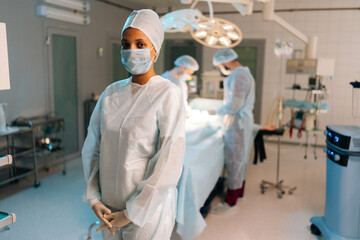 Portrait of tired African-American female doctor standing posing looking at camera in light operating theater. Diverse team of surgeons performing operation on background. Concept of emergency.