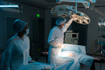 Male plastic surgeon in surgical uniforms and masks adjusting light before operation in dark operating theatre. Surgery, hospital, medical and healthcare services. Concept of surgery and emergency.