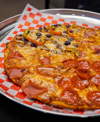 Sabrosa Pizza de Varios sabores, Pepperoni, Jamon, Queso, Champiñones, Aceitunas y Pimientos sobre papel de cuadros rojo y blanco