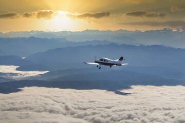 Kleinflugzeug fliegt über einer Wolkendecke in einen epischen Sonnenuntergang hinter einer Berglandschaft - obrazy, fototapety, plakaty