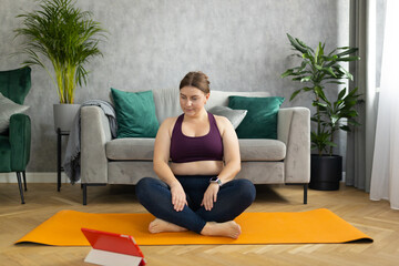 Sporty plus size woman in sportswear working out at home. Sports, activity and weight loss concept. Young plus size woman in leggings exercising on the mat, watching online video via tablet