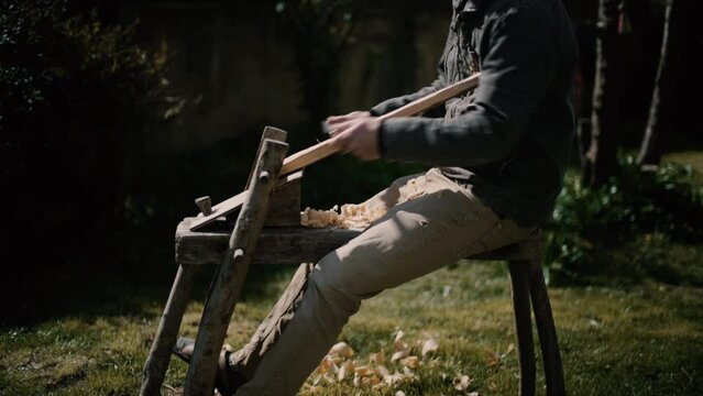 Carpenter using shaving horse for traditional carving, using draw knife, traditional tool carving wood into a cross, on sunny day on the grass