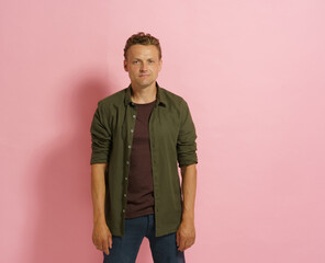 A man in a green shirt stands in front of a pink background. He looks serious and is wearing a brown shirt
