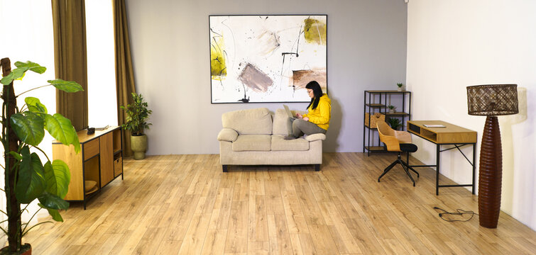 A woman is sitting on a couch in a living room with a painting on the wall. The room is decorated with a few potted plants and a lamp. The woman is reading a book, and the room has a cozy