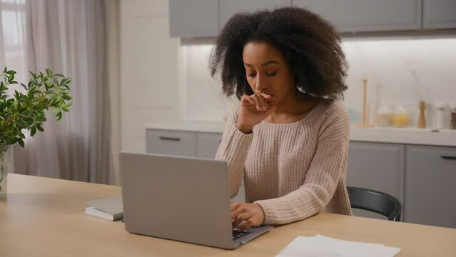 African American woman freelancer online working from home with laptop in kitchen young ethnic girl female businesswoman writer pensive thinking create idea typing article on computer distant studying