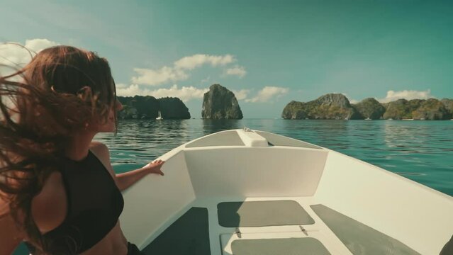 A woman wearing a black top is standing on a boat cruising through the waters of El Nido Islands in the Philippines. The scene captures her in motion, enjoying a boat tour experience.