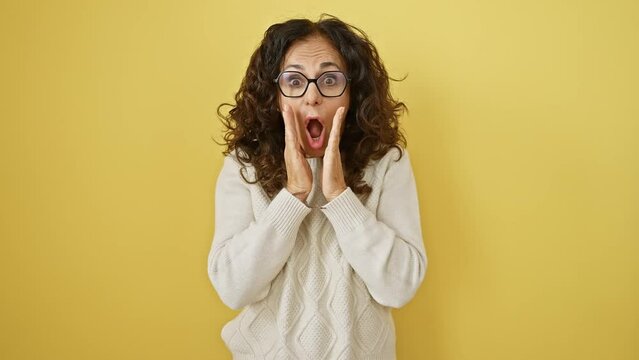 Middle age hispanic woman wearing glasses standing afraid and shocked, surprise and amazed expression with hands on face over isolated yellow background