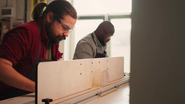 Woodworking expert wears protection gear, inserting plank in spindle moulder next to coworker. CNC machinist uses safety glasses while cutting lumber with wood shaper helped by colleague, camera B