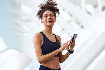 A woman in a sports bra is smiling while holding a cell phone standing outdoors - obrazy, fototapety, plakaty