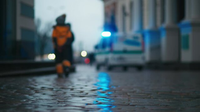 A figure walking on a wet city street next to an electric blue ambulance