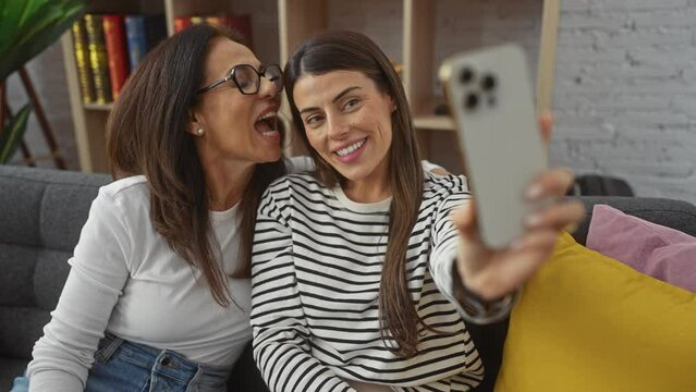 A mother and daughter in a cozy living room share a joyful moment taking a selfie together, radiating love and happiness.