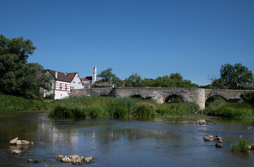 A historic stone bridge spans the river Wörnitz. The bridge from the Middle Ages is located in Harburg, Bavaria. The blue sky is reflected in the water.
