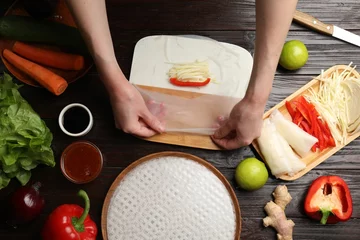 Foto op Canvas Making delicious spring rolls. Woman wrapping fresh vegetables into rice paper at wooden table, flat lay © New Africa