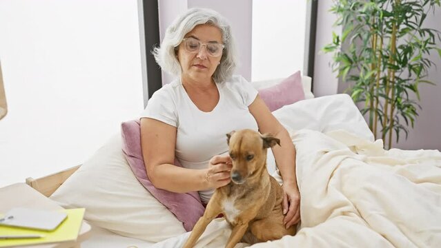 Confident middle age woman in pyjamas, pondering with a sober expression on her bed, grey-haired dog at her side in the bedroom