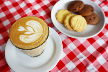 Choco chip cookies and latte coffee on a white plate with a checkered tablecloth seen above