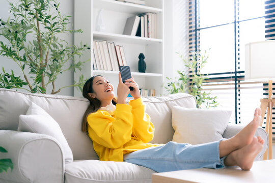 Happy young asian woman  wearing headset and relaxing on comfortable couch at home, texting messaging on smartphone, smiling girl use cellphone chatting, online shopping at home, video call