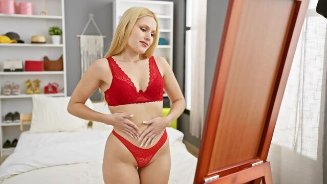 A young, attractive caucasian woman in red lingerie examines herself in a bedroom mirror, reflecting a cozy, lived-in apartment space.