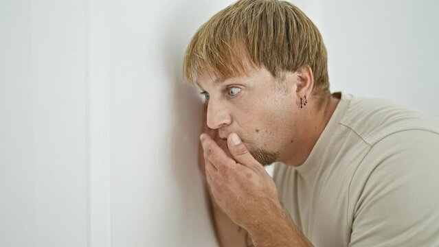 Young caucasian man with beard and blond hair eavesdropping at home, showing curiosity and anticipation.